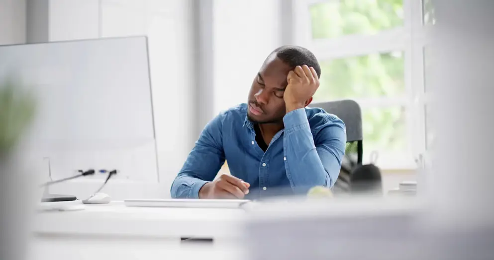 Man in a blue shirt sitting at a desk with head resting on hand, appearing fatigued. 