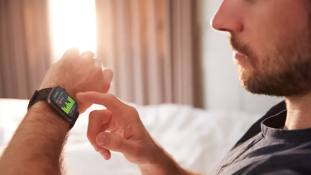 Man Sitting Up In Bed Looking At Screen Of Smart Watch