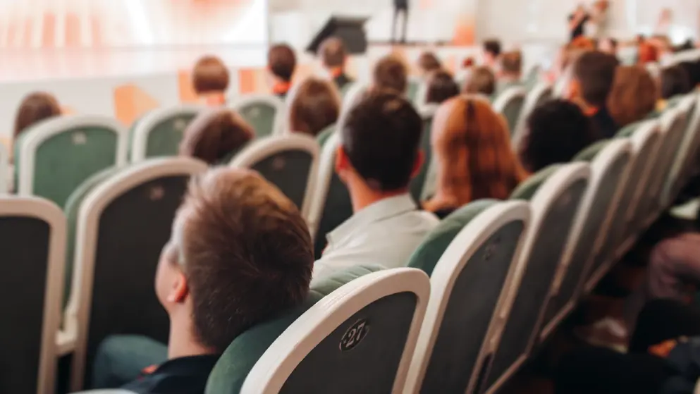 Audience at a modern conference hall listens to panel discussion, various audience members, green chairs