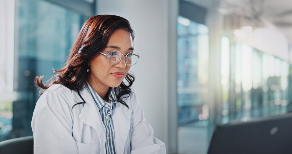 A female healthcare professional, with brown hair and wearing glasses, looks at her laptop in front of her.