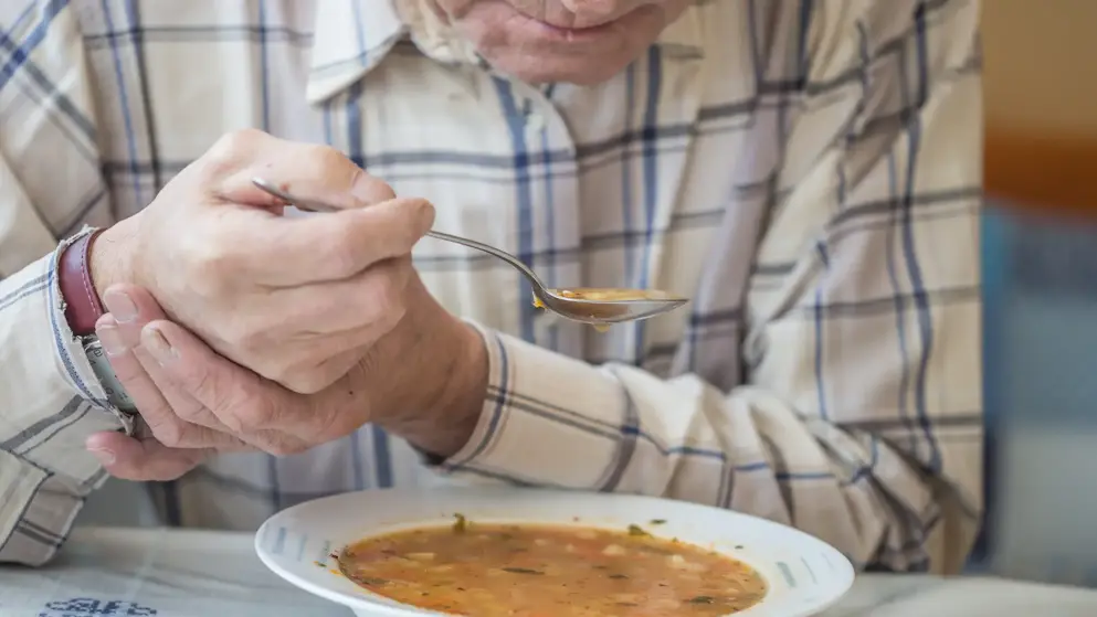 Elderly man struggles to lift soup to his mouth due to muscle tremors