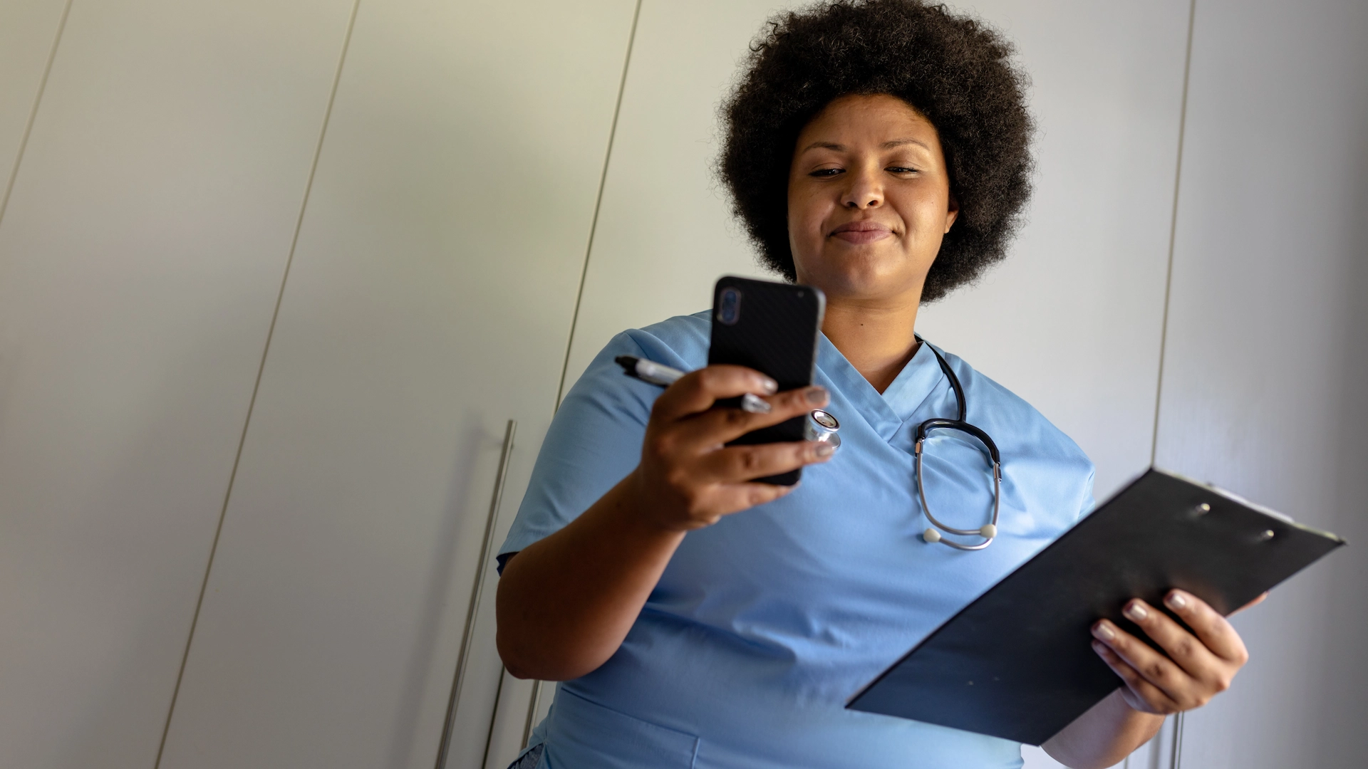 African american mid adult female nurse using smart phone while holding clipboard in hospital